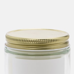 May include: A clear glass jar with a gold-colored metal lid. The lid has a ridged edge and a flat top. A white label is partially visible on the jar's side. The jar appears to be empty or filled with a white substance.