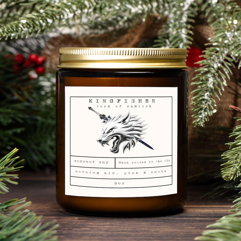 May include: A brown glass jar candle with a gold lid. The label features the text "KINGFISHER lord of cahlish" and a wolf illustration. The candle is scented with coconut soy and morning air, pine & smoke. It weighs 9 oz.