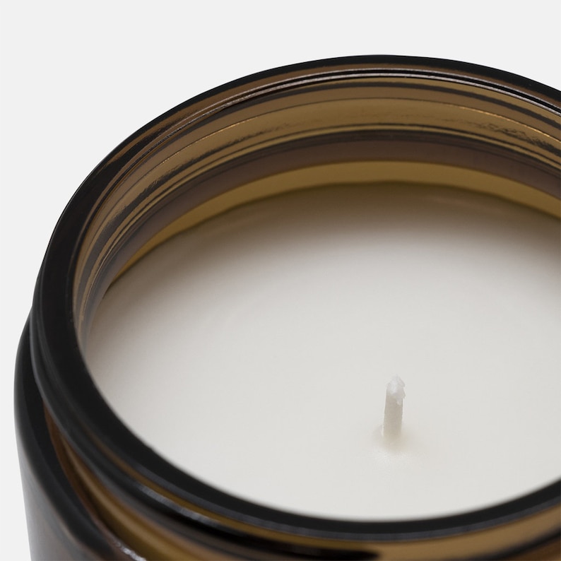 May include: Close-up of a brown glass jar candle with a white wax interior and a single white wick. The jar has a dark brown rim and a light brown body. The candle is set against a white background.