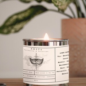 Puede incluir: Una lata de vela plateada con una etiqueta blanca que dice "NESTA Valkerie & Christian" y tiene una ilustraci&oacute;n en blanco y negro de una m&aacute;scara y una daga. La etiqueta tambi&eacute;n enumera las notas de aroma: Top: Eucaly Ozone, Middle: Sugar Marshmall, Base: Oak Patchouli, Smoke Embers. La vela est&aacute; encendida y la llama es visible.