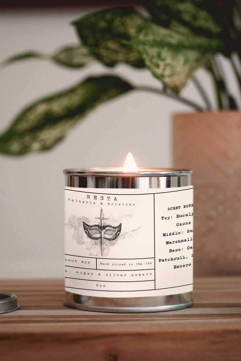 Puede incluir: Una lata de vela plateada con una etiqueta blanca que dice "NESTA Valkerie & Christian" y tiene una ilustraci&oacute;n en blanco y negro de una m&aacute;scara y una daga. La etiqueta tambi&eacute;n enumera las notas de aroma: Top: Eucaly Ozone, Middle: Sugar Marshmall, Base: Oak Patchouli, Smoke Embers. La vela est&aacute; encendida y la llama es visible.