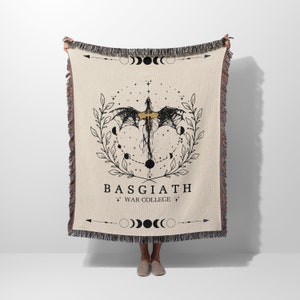 May include: A woven tapestry throw blanket with a beige background featuring a black and gold illustration of a dragon with wings spread, surrounded by a wreath of leaves and a crescent moon design. The text "BASGIATH" and "WAR COLLEGE" is printed in black below the dragon.