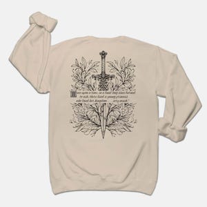 May include: A beige long-sleeve sweatshirt with a black and white illustration of a sword surrounded by floral designs. The text "once upon a time, in a land long since burned to ash, there lived a young princess who loved her kingdom... very much." is printed on the sweatshirt.