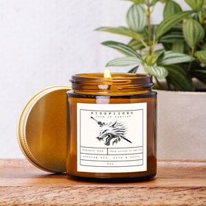 May include: A lit, amber-colored glass jar candle with a gold lid. The label reads "KINGFISHER lord of eahligh" with a wolf illustration. The candle is labeled "coconut soy" and "morning air, pine & smoke" with a 4 oz size.