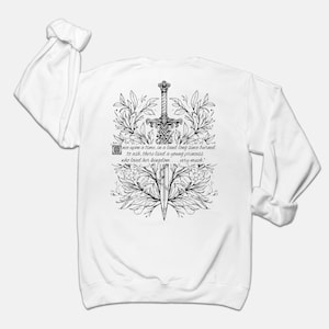 May include: White long-sleeve sweatshirt with a black and white illustration of a sword surrounded by floral designs. The text "once upon a time, in a land long since burned to ash, there lived a young princess who loved her kingdom... very much." is printed below the illustration.