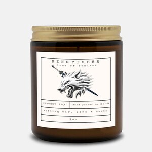 May include: A brown glass jar candle with a gold lid. The label features the text "KINGFISHER lord of eahlish" above a silver wolf head with a sword. Below, it reads "coconut soy, morning air, pine & smoke" and "9 oz."