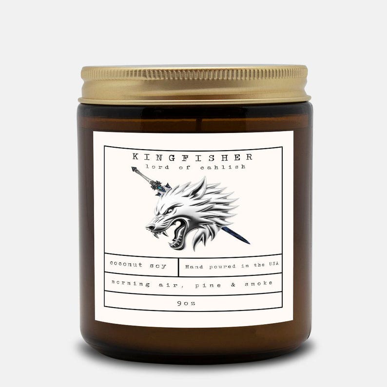 May include: A brown glass jar candle with a gold lid. The label features the text "KINGFISHER lord of eahlish" above a silver wolf head with a sword. Below, it reads "coconut soy, morning air, pine & smoke" and "9 oz."