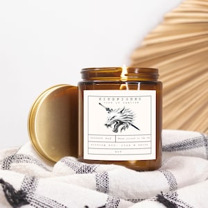 May include: A lit brown glass jar candle with a gold lid, labeled "KINGFISHER lord of ashlish". The label features a wolf graphic. The candle is on a black and white patterned cloth. The candle is 9 oz.