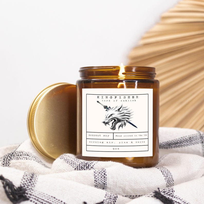 May include: A lit brown glass jar candle with a gold lid, labeled "KINGFISHER lord of ashlish". The label features a wolf graphic. The candle is on a black and white patterned cloth. The candle is 9 oz.