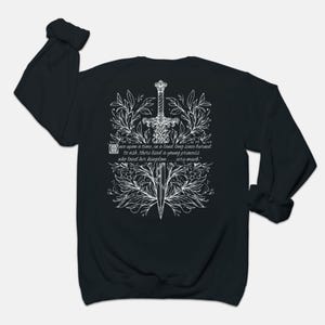 May include: Black long-sleeve sweatshirt with a white graphic design of a sword surrounded by flowers and leaves. The text "once upon a time, in a land long since burned to ash, there lived a young princess who loved her kingdom... very much." is printed on the sweatshirt.