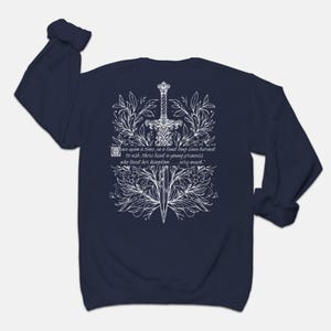 May include: Navy blue long-sleeve sweatshirt with a white design of a sword surrounded by flowers and leaves. The text "once upon a time, in a land long since burned to ash, there lived a young princess who loved her kingdom... very much." is printed on the sweatshirt.