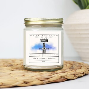 May include: A glass jar candle with a gold lid, featuring a label with the text "Dorian Havilliard King of Adarlan." The label includes a sword and crown design, with the scent "sea & winter morning." The candle is 9 oz.