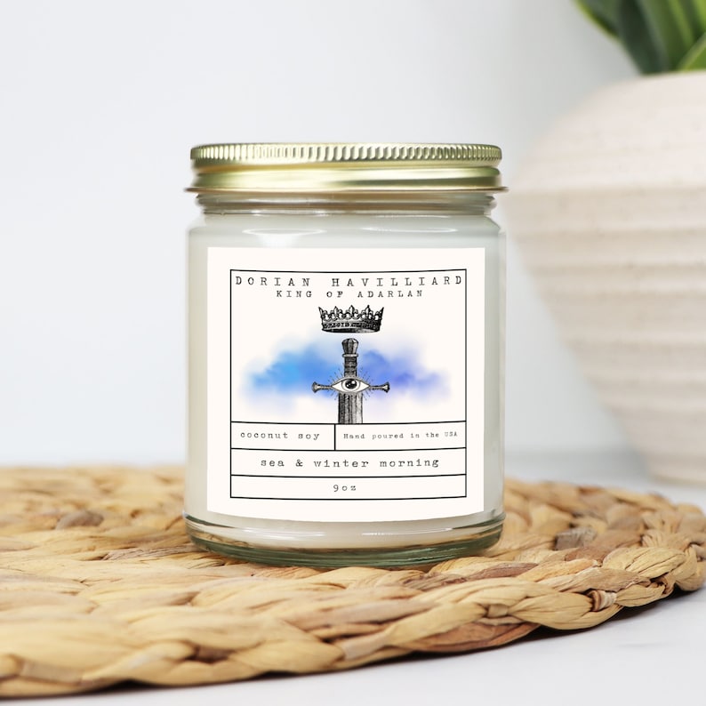 May include: A glass jar candle with a gold lid, featuring a label with the text "Dorian Havilliard King of Adarlan." The label includes a sword and crown design, with the scent "sea & winter morning." The candle is 9 oz.