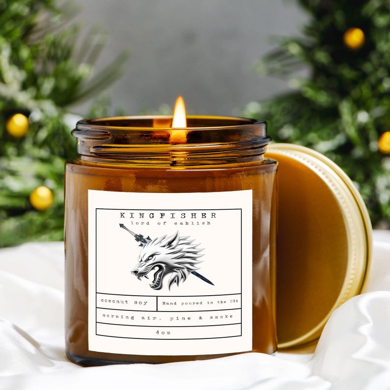 May include: A lit, amber-colored candle in a glass jar with a gold lid. The label reads "KINGFISHER lord of oahlish" with a wolf illustration. The candle is made with coconut soy and has a scent of morning air, pine, and smoke. It weighs 4 oz.
