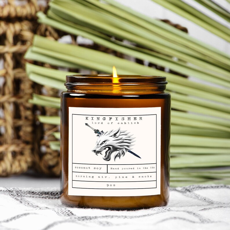 May include: A lit, brown glass jar candle with a label that reads "KINGFISHER lord of cahlish." The label features a wolf illustration and the text "coconut soy, morning air, pine & smoke, 9oz."