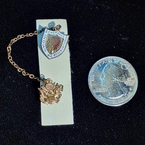 May include: A vintage gold-toned pin set featuring a shield-shaped emblem with the text "CHEROKEE & N.C. RESERVATION" and an image of a Native American. The set includes a smaller emblem and a chain, next to a US quarter.