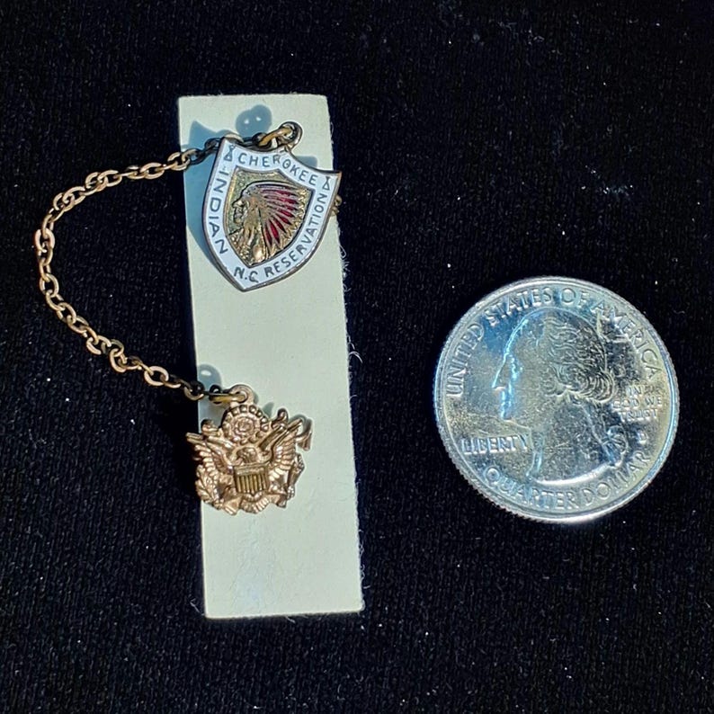 May include: A vintage gold-toned pin set featuring a shield-shaped emblem with the text "CHEROKEE & N.C. RESERVATION" and an image of a Native American. The set includes a smaller emblem and a chain, next to a US quarter.