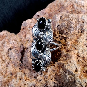 May include: A silver ring featuring three vertically aligned, oval-shaped black stones set in serrated bezels. The metal band is decorated with embossed leaf patterns and small silver spheres between the stones, creating a textured, elongated design.