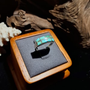 May include: A silver ring with a rectangular turquoise inlay. The turquoise is arranged in a grid pattern across the top of the ring. The ring is displayed on a wooden jewelry box with a dark gray interior.
