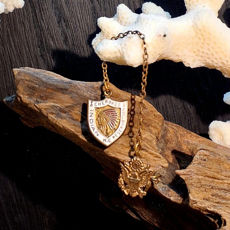 May include: A gold-toned pendant necklace featuring a shield-shaped charm with the text "INDIAN CHEROKEE N.C. RESERVATION" and an image of a Native American. A smaller charm with an eagle emblem hangs below.