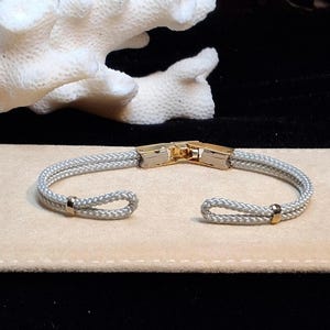 May include: A silver-toned braided cord bracelet featuring a gold-toned metal clasp at the center and two small gold-toned metal accents near the open ends. The bracelet is constructed from two parallel strands of cord that form loops at each terminal.