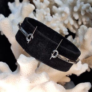 May include: A black bracelet with a silver clasp. The bracelet is made of a soft, dark material and features a geometric design on the clasp. The bracelet is displayed on a white coral background.