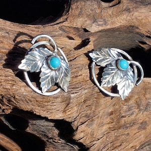 May include: A pair of silver-toned leaf-shaped earrings with a turquoise stone center. The earrings have a detailed leaf design and are set within a circular frame. The earrings are displayed on a textured wooden surface.