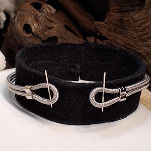 May include: A silver-toned bracelet with a woven design and a unique clasp. The bracelet is displayed on a black velvet surface, showcasing its intricate details and craftsmanship. The jewelry is a stylish accessory.
