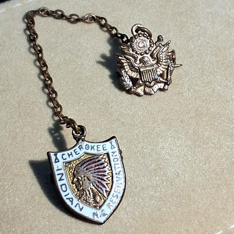 May include: Vintage pin set with a chain. One pin features a shield with "CHEROKEE INDIAN RESERVATION" text and a Native American profile. The other pin is a gold-colored eagle emblem. The pins are connected by a chain.