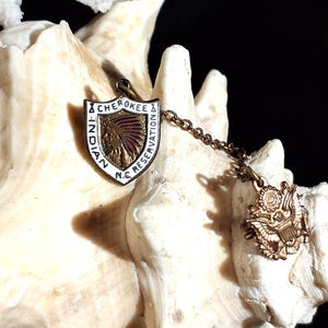 May include: Vintage shield-shaped pin with the text "CHEROKEE INDIAN N.C. RESERVATION". The pin features a Native American headdress design in gold and red. A smaller gold-colored emblem with an eagle is attached by a chain.