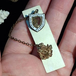 May include: A vintage gold-toned pin set featuring a shield-shaped emblem with the text "Cherokee Indian Reservation N.C." and an image of a Native American chief. The set includes a smaller emblem with an eagle design, connected by a delicate chain.