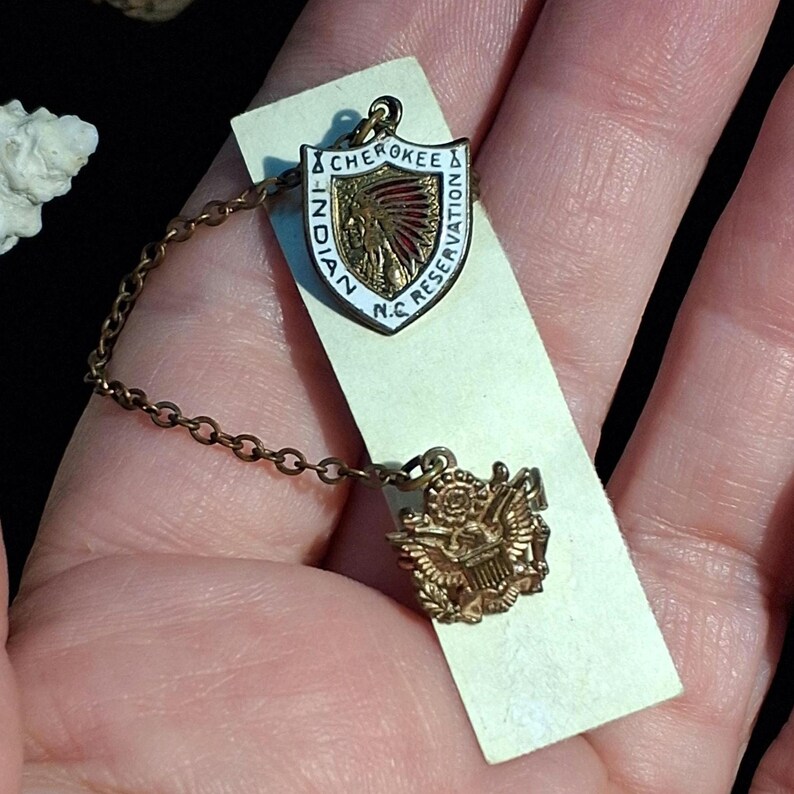 May include: A vintage gold-toned pin set featuring a shield-shaped emblem with the text "Cherokee Indian Reservation N.C." and an image of a Native American chief. The set includes a smaller emblem with an eagle design, connected by a delicate chain.