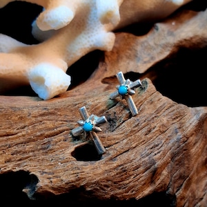 May include: A pair of silver cross stud earrings with a small turquoise stone in the center of each cross. The earrings are displayed on a piece of weathered wood, with a white coral-like structure in the background.