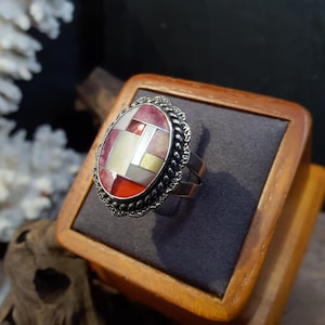 May include: Oval-shaped silver ring with a mosaic design. The ring features a geometric pattern of red, white, and yellow-toned rectangular shapes. The ring is set on a wooden display box with a gray interior.
