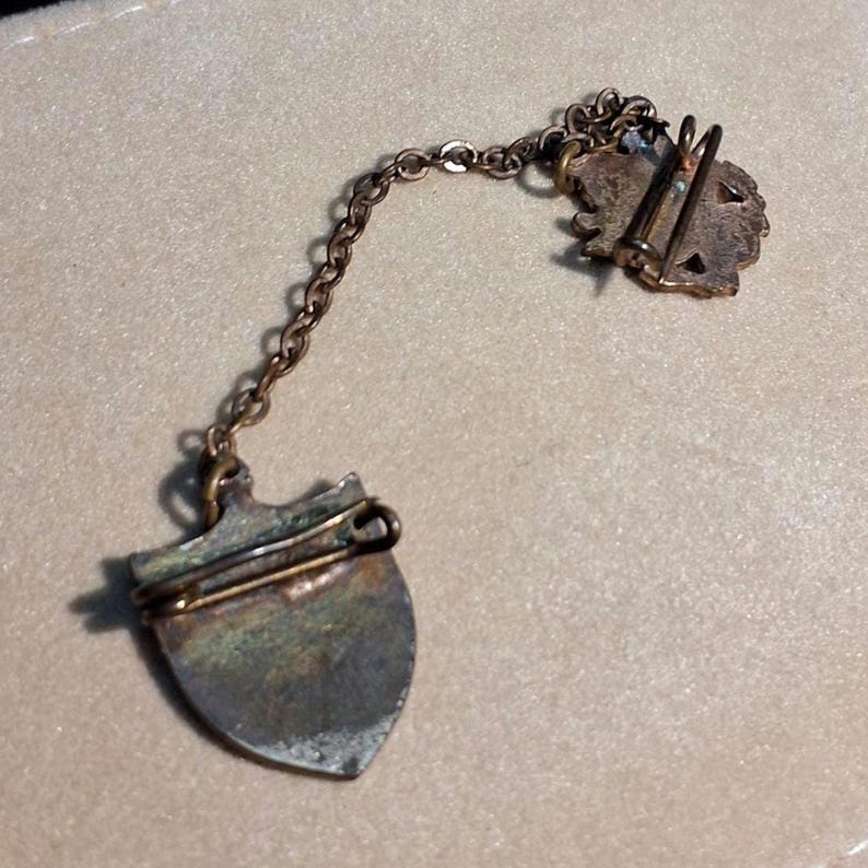 May include: Vintage silver-toned metal pin with a shield-shaped pendant and a decorative floral element. The pin is connected to the pendant by a delicate chain. The pin has a clasp on the back for secure attachment.