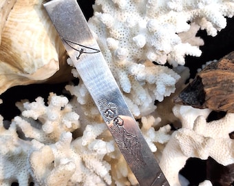 May include: A silver letter opener with a turquoise stone at the top. The opener has an etched design of a bird and feathers. The background includes white coral and a seashell.
