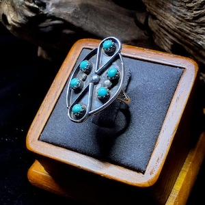 May include: A silver ring with an oval design, featuring six turquoise stones and small silver accents. The ring is displayed on a wooden jewelry box with a dark gray interior, showcasing the ring's intricate details.