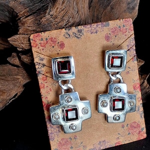 May include: A pair of silver drop earrings featuring a square-cut deep red garnet stone at the top and a larger cross-shaped pendant below. The cross section contains a central square garnet and four small white pearls set in polished silver.