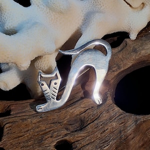 May include: A silver-toned cat brooch in a playful pose, with its tail arched and body curved. The brooch has a detailed head and is set against a natural wood background, evoking a sense of artistry and charm. The cat's design is simple yet elegant.