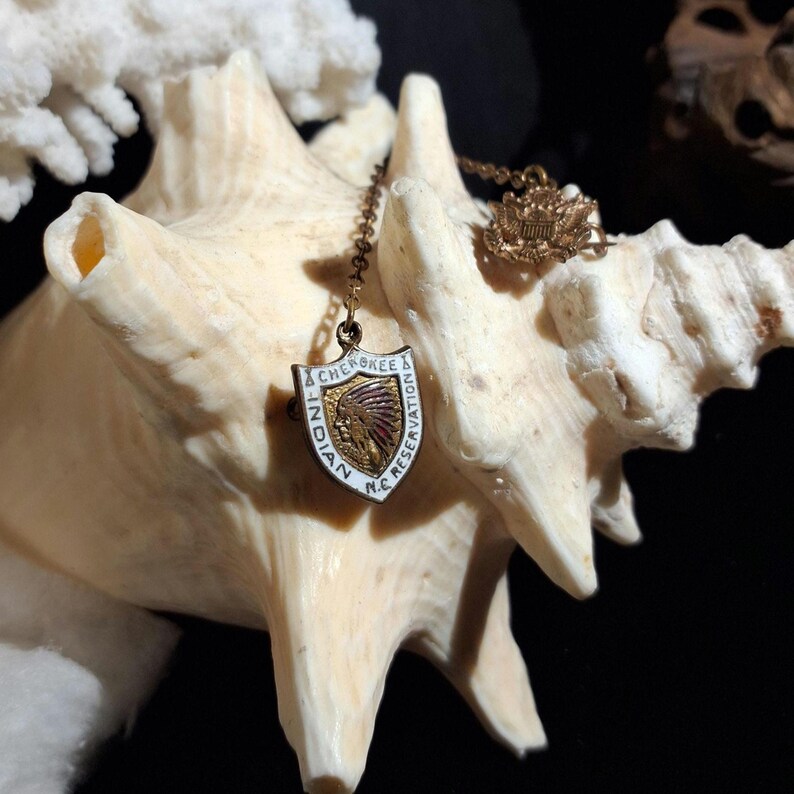 May include: A vintage Cherokee Indian Reservation pendant with a shield design. The pendant features a Native American headdress in red and gold, with the words "Cherokee Indian Reservation N.C." in a white and gold border. The pendant hangs from a delicate chain.