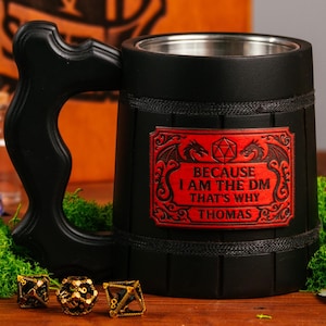 以下が含まれることがあります： 「BECAUSE I AM THE DM THAT'S WHY THOMAS」と書かれた赤いラベルが付いた、ハンドル付きの黒いマグカップ。 マグカップは、黒いドクロ、サイコロ、小さなガラス瓶が置かれた木製のテーブルの上にあります。 D&Dのロゴが入った木箱が背景にあります。