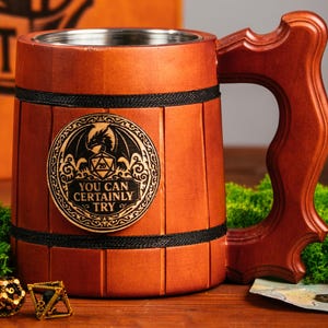 May include: A wooden tankard with a stainless steel interior and a decorative handle. The tankard features a metal emblem with a dragon and the text "YOU CAN CERTAINLY TRY". A wooden box with the text "WELL, SHIT" is in the background.
