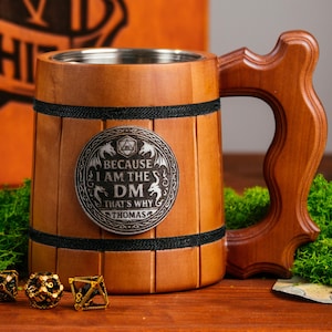 Könnte beinhalten: Holzkrug mit Edelstahl-Innenraum und Holzgriff. Der Krug hat ein dekoratives Metallabzeichen mit der Aufschrift "BECAUSE I AM THE DM THAT'S WHY THOMAS". Im Hintergrund befinden sich Würfel, eine Holzkiste und ein Totenkopf.