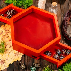 Peut inclure: Un plateau à dés hexagonal en bois rouge vif doté d'un tiroir de rangement coulissant. Le plateau présente une surface intérieure lisse et plate ainsi qu'une section séparée plus petite. Plusieurs dés polyédriques sont visibles à l'intérieur.