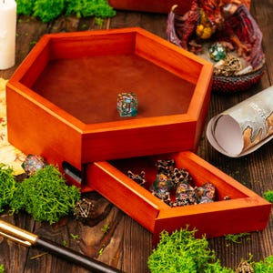 Puede incluir: Una bandeja hexagonal de madera para dados con un interior forrado en cuero marrón y un cajón extraíble. La bandeja contiene un dado translúcido multicolor, mientras que el cajón alberga varios dados adicionales.