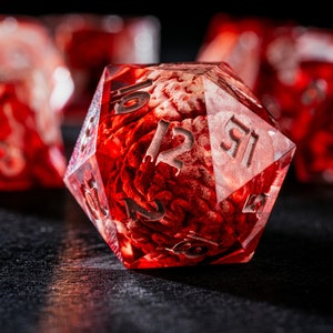 Puede incluir: Un dado de veinte caras rojo transl&uacute;cido que contiene un modelo anat&oacute;mico de cerebro incrustado en la resina. El dado tiene bordes afilados y n&uacute;meros grabados en color plateado. Otros dados rojos similares son visibles en el fondo borroso.