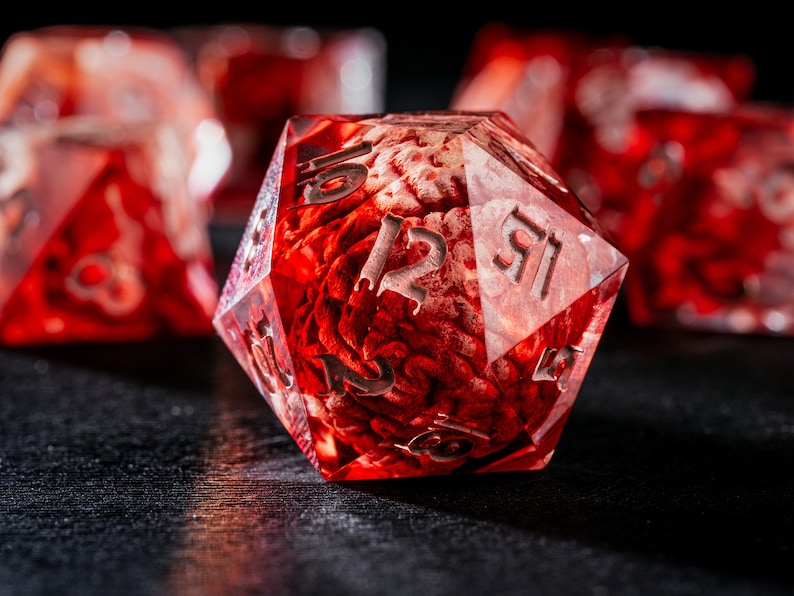 Puede incluir: Un dado de veinte caras rojo transl&uacute;cido que contiene un modelo anat&oacute;mico de cerebro incrustado en la resina. El dado tiene bordes afilados y n&uacute;meros grabados en color plateado. Otros dados rojos similares son visibles en el fondo borroso.