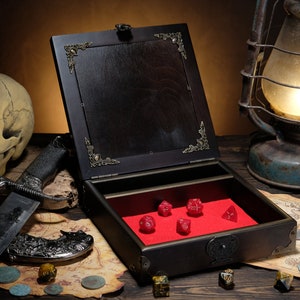 May include: A dark wooden box with an open lid featuring ornate metal corner accents and a bright red velvet interior lining. Inside the box are five red polyhedral dice. The box is positioned on a map with scattered coins and a large knife.