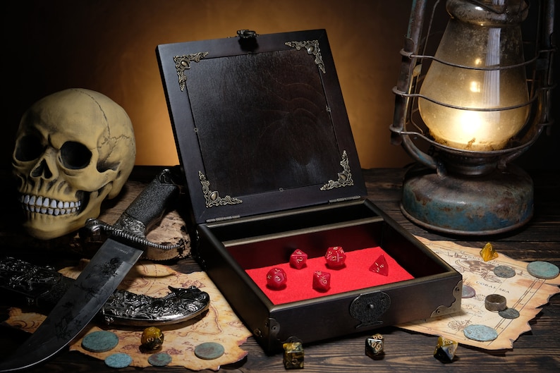 May include: A dark wooden box with an open lid featuring ornate metal corner accents and a bright red velvet interior lining. Inside the box are five red polyhedral dice. The box is positioned on a map with scattered coins and a large knife.
