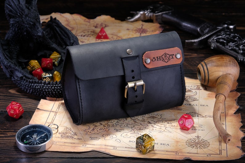 May include: A matte black leather pouch with a front buckle closure and a brown leather patch featuring the text SHOT and a geometric icon. The pouch is constructed with visible stitching along the edges and secured with metallic rivets and a buckle.